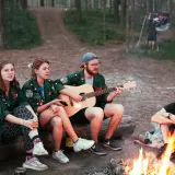 Grupa harcerzy siedzi przy ognisku, jeden z nich gra na gitarze. W tle widać las i flagę. Wspólne śpiewanie piosenek harcerskich przy ognisku.