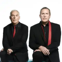Andy McCluskey i Paul Humphreys z OMD, duet znany z niezapomnianych piosenek, w czarnych garniturach i czerwonych akcentach.