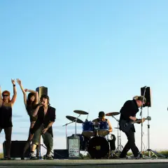 Zespół rockowy na tle nieba. Wokalista, gitarzysta, basista, perkusista i dwie tancerki. Zespoły rockowe zagraniczne alfabetycznie.