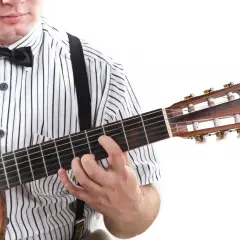 Dłoń na gryfie gitary, palce układają akord. W tle widać fragment koszuli w paski i muszkę. To jak czytać tabulatura gitarowa oznaczenia.