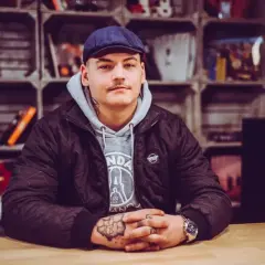 Młody mężczyzna w czapce i kurtce, z tatuażami na rękach, siedzi przy stole. W tle regały z płytami i innymi przedmiotami. To jego **frosti wiek**.