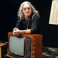 Mężczyzna z długimi siwymi włosami i okularami opiera się o stary telewizor. Wokół niego lampy i inne telewizory. Widać, że trzeba wiedzieć, kiedy ze sceny zejść.