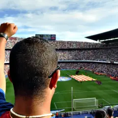 Widok z trybun stadionu na murawę, gdzie kibice tworzą mozaikę z flag, a w tle słychać hymn Barcelony.
