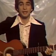 Mężczyzna w garniturze i wzorzystej koszuli gra na gitarze. W tle widać materiał, na którym **ciągle pada tekst**.