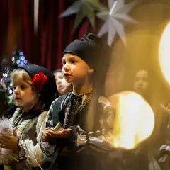 Dzieci w tradycyjnych strojach śpiewają kolędy. W tle choinka i gwiazdy.