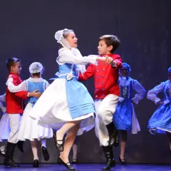 Dzieci w strojach krakowiaków tańczą na scenie. Chłopiec w czerwonym żupanie i dziewczynka w błękitnej sukience wirują w tańcu.