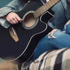 Osoba w dżinsowej kurtce i podartych jeansach gra na czarnej gitarze akustycznej. Słychać dźwięki muzyki.