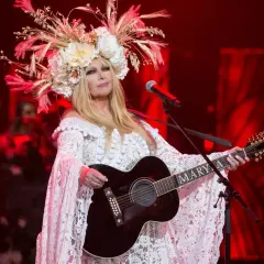 Artystka w białej, koronkowej sukni i kwiecistym wianku gra na gitarze. Jej muzyka sprawia, że pokonamy fale wykonawcy.