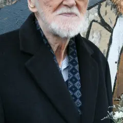 Starszy pan z siwą brodą, elegancko ubrany w czarny płaszcz i wzorzystą apaszkę, to legendarny walicki muzyk jazzowy.
