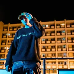 Młoda dziewczyna w czapce i swetrze z napisem, stoi na tle bloku mieszkalnego nocą.