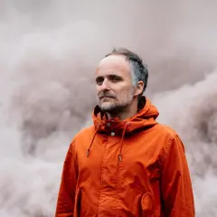 Artur Rojek w pomarańczowej kurtce, na tle dymu. Jego utwory niosą emocje.