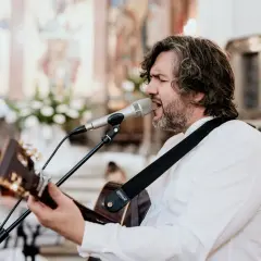 Mężczyzna z gitarą śpiewa pieśni chrześcijańskie w kościele. W tle widać ołtarz i obrazy.
