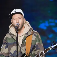 Artysta w czapce z daszkiem i kurtce moro śpiewa do mikrofonu, trzymając gitarę. Może to jego żona czeka na niego po koncercie?