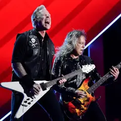 Dwóch gitarzystów zespołu Metallica gra na scenie na tle czerwonej kurtyny.