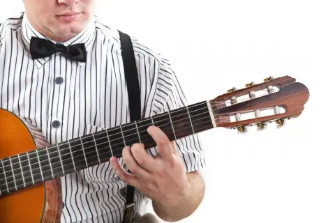 Dłoń na gryfie gitary, palce układają akord. W tle widać fragment koszuli w paski i muszkę. To jak czytać tabulatura gitarowa oznaczenia.