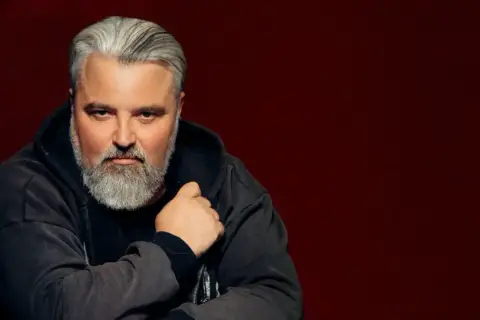 Mirosław Niewiadomski, z siwą brodą i włosami, w bluzie z kapturem. Jego życiorys to historia pasji i determinacji.