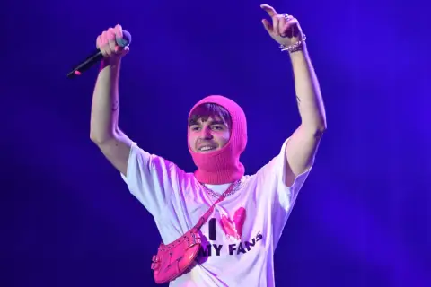Artysta w różowej kominiarce i koszulce "I ❤️ MY FANS" z torebką na ramię, z mikrofonem w dłoni, pozdrawia publiczność.