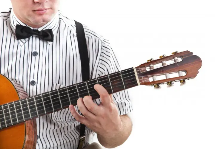 Dłoń na gryfie gitary, palce układają akord. W tle widać fragment koszuli w paski i muszkę. To jak czytać tabulatura gitarowa oznaczenia.