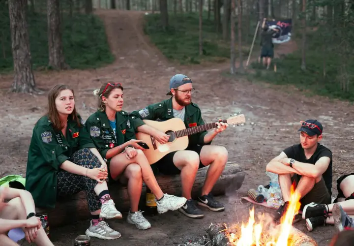 Grupa harcerzy siedzi przy ognisku, jeden z nich gra na gitarze. W tle widać las i flagę. Wspólne śpiewanie piosenek harcerskich przy ognisku.