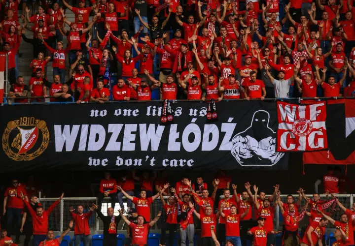 Tłum kibiców Widzewa w czerwonych koszulkach skanduje przyspiewki na Widzew, machając rękami. Na banerze widnieje napis "WIDZEW ŁÓDŹ".