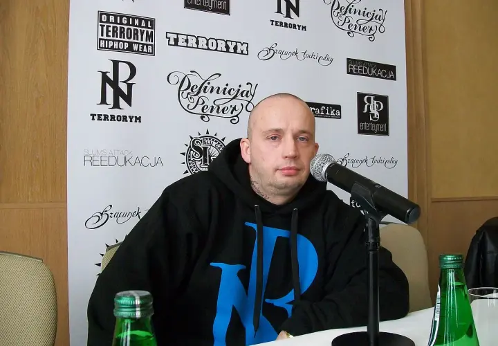 Peja w czarnej bluzie z niebieskim logo, przed mikrofonem. Na ścianie za nim napisy: "TERRORYM", "REEDUKACJA", "Definicja Peja". Wiecie co z nim zrobić.