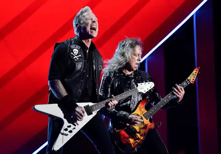 Dwóch gitarzystów zespołu Metallica gra na scenie na tle czerwonej kurtyny.