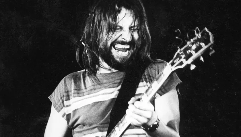 Rockman z długimi włosami i brodą, grający na gitarze z pasją. Jego twarz wyraża emocje, jakby to była jego osobista dżem modlitwa.