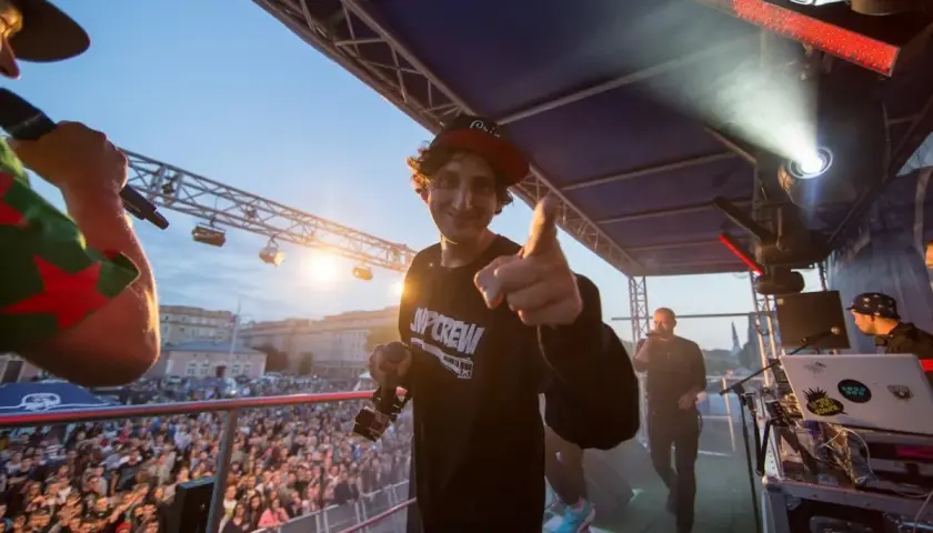 Kosi z JWP Crew w wieku około 30 lat, na scenie z mikrofonem, wskazuje palcem na widownię.