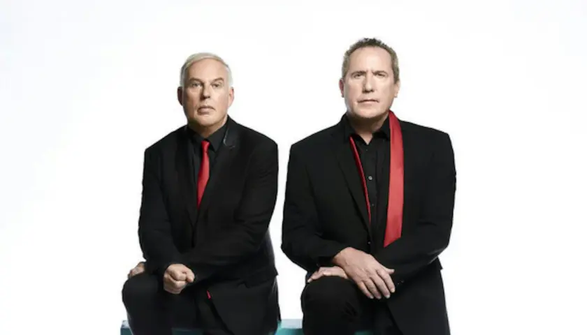 Andy McCluskey i Paul Humphreys z OMD, duet znany z niezapomnianych piosenek, w czarnych garniturach i czerwonych akcentach.