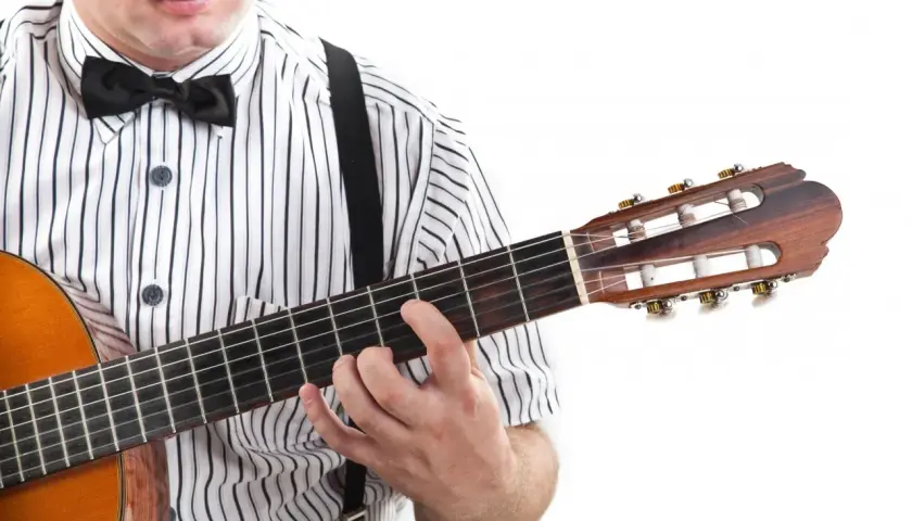 Dłoń na gryfie gitary, palce układają akord. W tle widać fragment koszuli w paski i muszkę. To jak czytać tabulatura gitarowa oznaczenia.