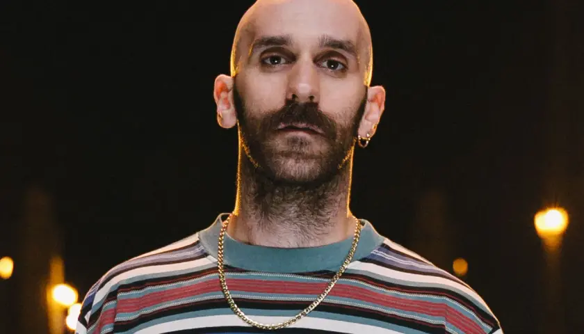 Łysy mężczyzna z zarostem, w kolorowej bluzie i złotym łańcuszku, wygląda jakby miał zaraz zacząć śpiewać "x ambassadors unsteady tekst".