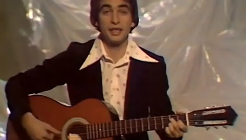 Mężczyzna w garniturze i wzorzystej koszuli gra na gitarze. W tle widać materiał, na którym **ciągle pada tekst**.