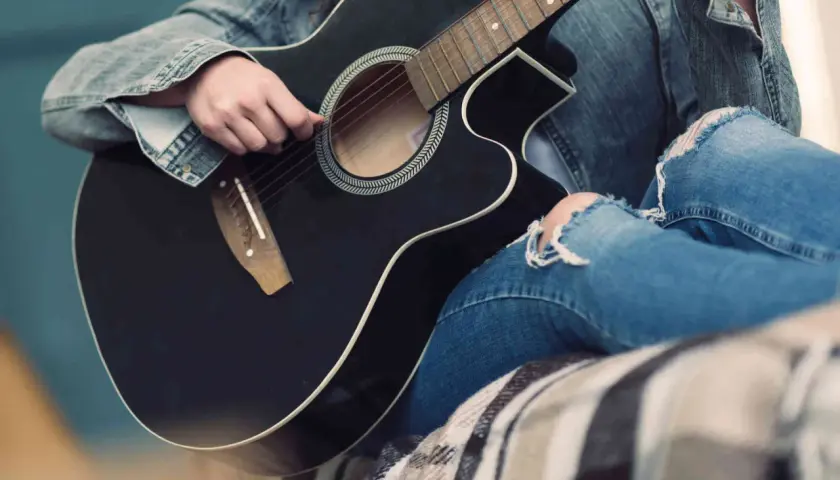 Osoba w dżinsowej kurtce i podartych jeansach gra na czarnej gitarze akustycznej. Słychać dźwięki muzyki.