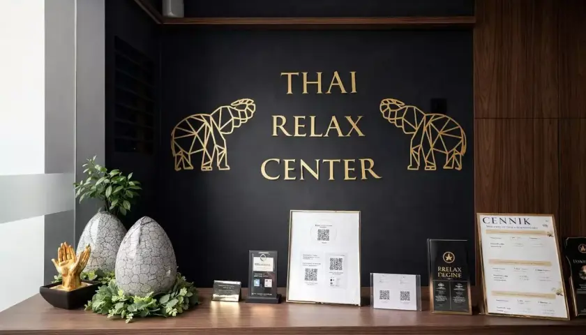 Recepcja centrum masażu tajskiego. Złote słonie i napis "THAI RELAX CENTER" na czarnej ścianie. Na blacie dekoracje i cennik. Tu możesz się wyciszyć.