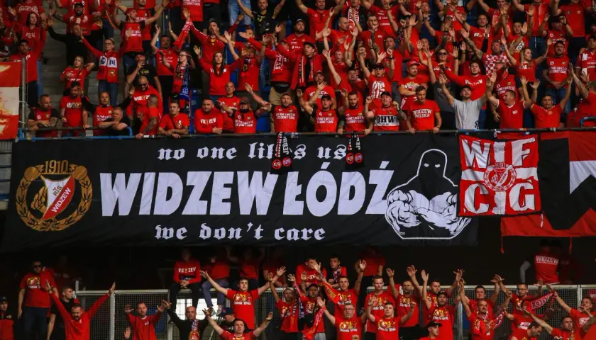 Tłum kibiców Widzewa w czerwonych koszulkach skanduje przyspiewki na Widzew, machając rękami. Na banerze widnieje napis "WIDZEW ŁÓDŹ".