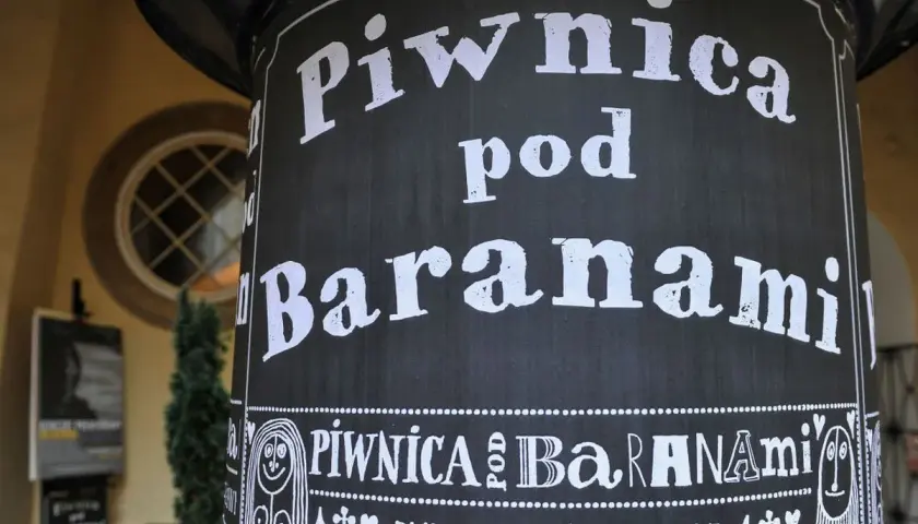 Czarny słup z białym napisem "Piwnica pod Baranami" i ozdobnymi elementami.