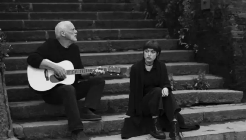 David Gilmour gra na gitarze, a obok niego siedzi jego córka.