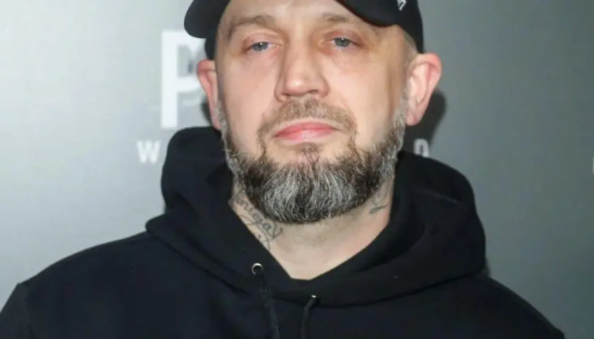Mężczyzna w czarnej bluzie z kapturem i czapce z daszkiem, z brodą i tatuażami na szyi. Pikers ile ma lat?