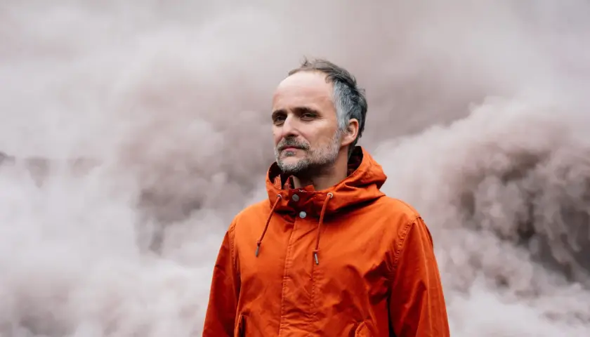 Artur Rojek w pomarańczowej kurtce, na tle dymu. Jego utwory niosą emocje.