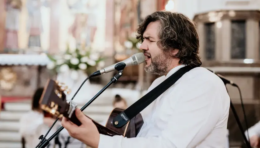 Mężczyzna z gitarą śpiewa pieśni chrześcijańskie w kościele. W tle widać ołtarz i obrazy.