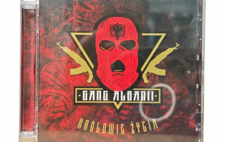 Okładka płyty CD "Królowie życia" zespołu Gang Albanii. Czerwona kominiarka z orłem, złote kałasznikowy i mroczne tło.