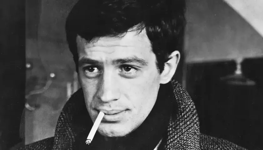Jean-Paul Belmondo z papierosem w ustach, w płaszczu. Jego spojrzenie przywodzi na myśl niezapomniane **belmondo cytaty**.