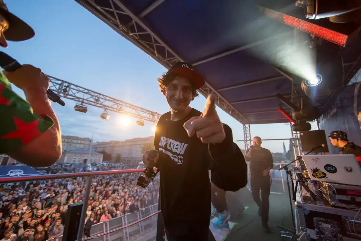 Kosi z JWP Crew w wieku około 30 lat, na scenie z mikrofonem, wskazuje palcem na widownię.