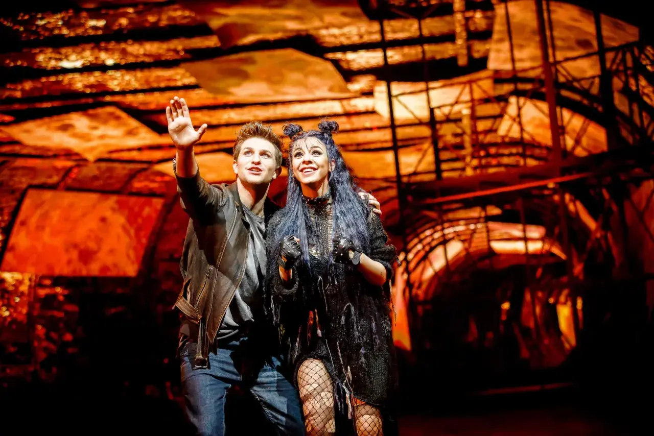 Para z musicalu "We Will Rock You" w polskim tłumaczeniu, pozdrawia publiczność na tle scenografii.