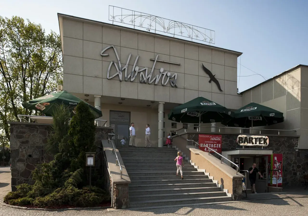 Budynek z napisem "Albatros" i sylwetką ptaka. Na schodach widać siedem dziewcząt z albatrosa, bawiących się w upalny dzień.