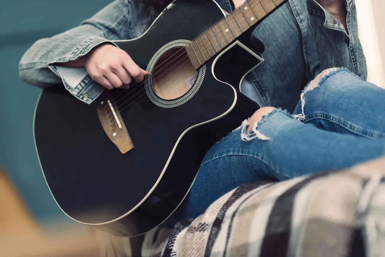 Osoba w dżinsowej kurtce i podartych jeansach gra na czarnej gitarze akustycznej. Słychać dźwięki muzyki.