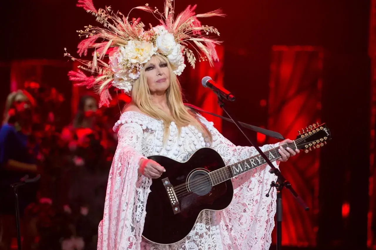 Artystka w białej, koronkowej sukni i kwiecistym wianku gra na gitarze. Jej muzyka sprawia, że pokonamy fale wykonawcy.