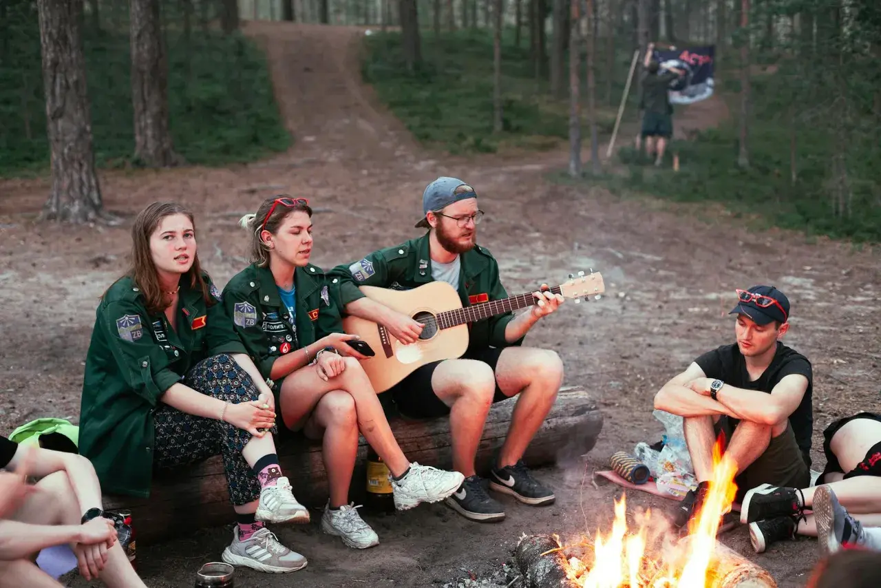 Grupa harcerzy siedzi przy ognisku, jeden z nich gra na gitarze. W tle widać las i flagę. Wspólne śpiewanie piosenek harcerskich przy ognisku.
