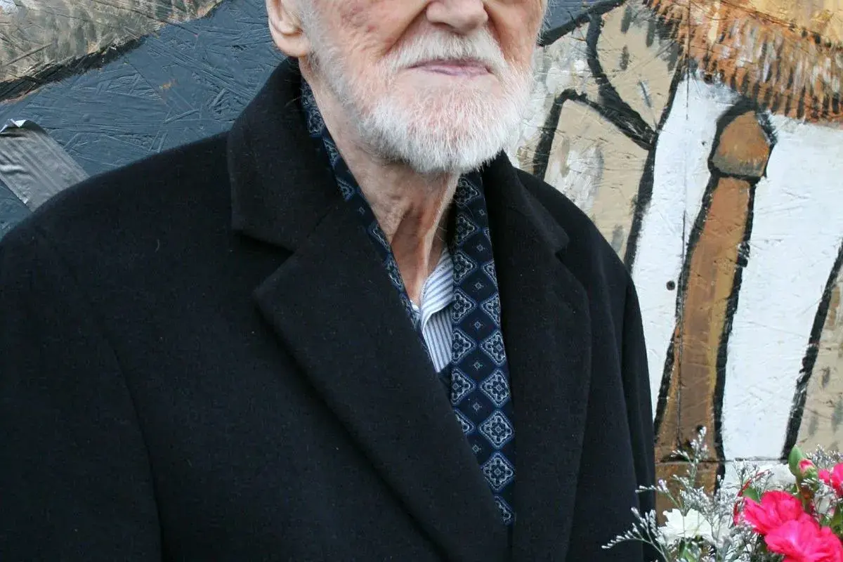 Starszy pan z siwą brodą, elegancko ubrany w czarny płaszcz i wzorzystą apaszkę, to legendarny walicki muzyk jazzowy.