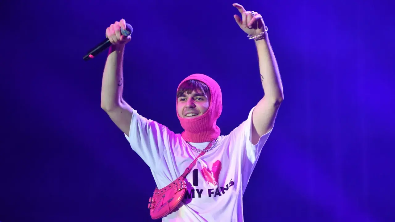 Artysta w różowej kominiarce i koszulce "I ❤️ MY FANS" z torebką na ramię, z mikrofonem w dłoni, pozdrawia publiczność.